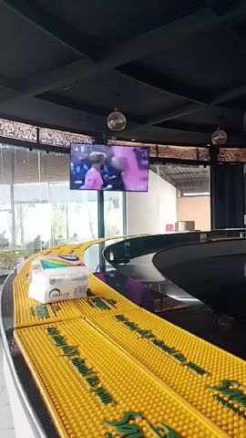 The video shows the inside of a bar. The camera pans from left to right, revealing a yellow bar top with a raised section in the center, a flat-screen TV showing a soccer game, and shelves filled with various liquor bottles. A box and a mixer are also visible on the shelves. A refrigerator stocked with drinks is seen to the right of the shelves, and a person in a white shirt briefly appears in the background. The overall setting suggests a casual bar environment.