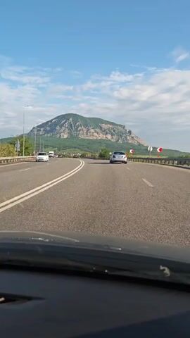 The video shows a view from inside a car driving on a multilane highway. The sky is blue with scattered clouds. In the distance, a large, flat-topped mountain covered in greenery dominates the horizon. The road has white lane markings and gray asphalt. Other cars are visible on the road. On the right side of the road, there's a metal guardrail and green vegetation. There are streetlights along the side of the highway.