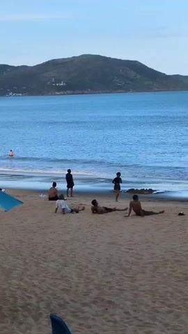 The video shows a beach scene with several people on the sand and in the water. In the foreground, a blue object, possibly part of a beach umbrella or toy, is visible. Further back on the sandy beach, a group of five individuals are present. Two are sitting, one leaning back on their arms. Another person is lying down on their stomach, while two others are reclined on their backs with their legs extended. A person in a dark shirt stands near the water's edge, facing away from the camera. Another person, also in dark clothing, is standing and walking towards the water. In the background, the blue ocean stretches to the horizon, with gentle waves lapping the shore. A distant, green, tree-covered hill forms the backdrop. One person is visible in the water, further out from the shore, appearing to be swimming. The sky is a pale blue with some scattered clouds. The overall atmosphere is relaxed, suggesting a leisurely day at the beach.