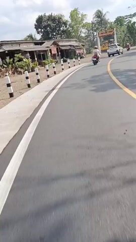 The video shows a view from a moving vehicle on a paved road. On the left side of the road are several wooden buildings with thatched roofs, and black and white striped bollards. A yellow road roller is visible behind a sign that reads "Warung Makan Sendang Sewu." Further down the road, a person on a motorcycle is riding ahead, followed by a white car. The road has a yellow line down the center. The environment appears to be rural with trees and vegetation. The sky is partly cloudy.