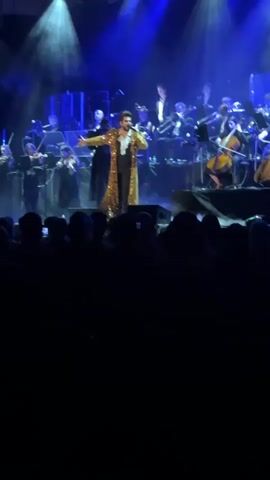 A person wearing a long gold sequined coat and dark pants stands on a stage, singing into a microphone. Behind them is an orchestra, consisting of string and brass instruments. The stage is bathed in blue and white spotlights. A crowd of people, mostly silhouetted, watches the performance from below the stage.