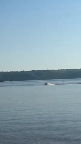A jet ski is being ridden across a calm body of water. The rider is visible as a small silhouette in the distance. The water is mostly still with slight ripples caused by the jet ski. There is a green, hilly landscape in the background. The sky is clear and blue.
