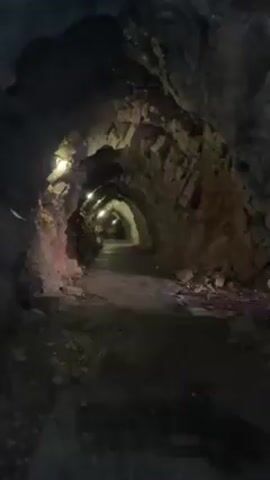 The video shows a slightly wet, paved path that goes through an arched tunnel. The tunnel is made of rock with lights above. The angle is from a first-person perspective while standing still.