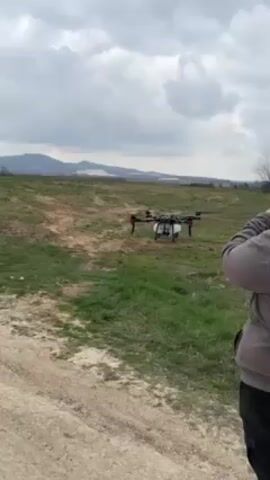 Here is a description of the video:

The video shows a large, black drone with eight propellers sitting on the ground in a grassy field. The drone has a white tank attached to its underside, and spray nozzles are visible underneath the tank. A person in a grey hoodie and black pants stands to the side of the drone, presumably controlling it. The background consists of a dirt road in the foreground, a green field with patches of dirt, and mountains in the distance under a cloudy sky. The drone's propellers begin to spin faster as the video progresses, suggesting it is preparing for takeoff.