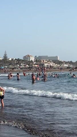 The video shows a crowded beach with many people in the water. Waves are breaking near the shore. In the background, there are buildings and palm trees. The sky is clear and blue.