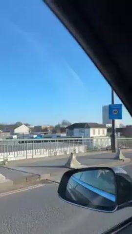 The video shows a driver’s perspective from inside a car, driving along a multi-lane road on a sunny day. The car is likely moving through a suburban or urban area, based on the presence of other cars, buildings, streetlights, and trees visible outside. A few pedestrians, including one with a bicycle, can be seen on the side of the road, waiting to cross the street. A building with a distinctive architecture can be seen in the distance. The car's interior is visible, featuring a black dashboard and trim, and the rear-view mirrors. Music can be heard playing in the background.