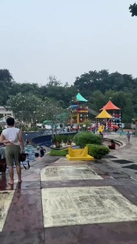 A vertical shot shows a man walking in an outdoor pool area, away from the camera, on a paved path. The man wears a white t-shirt and green shorts and carries a black bag over his left shoulder. To the left is a blue swimming pool with several people swimming. In the distance, a large water park area is visible with colorful slides and structures. The sky is overcast and grey. The ground in the foreground is tiled in a checkered pattern of light and dark squares, some of which are dirty.