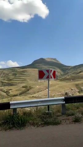 The video shows a mountainous landscape on a clear day. The foreground features a metal guardrail with black and white stripes. Beyond the guardrail, rolling hills are visible, some of which are terraced, suggesting agricultural use. A winding dirt path or road can be seen snaking up the hillside towards a prominent, rounded mountain peak in the distance. The sky is blue with scattered white clouds. The overall impression is a rural, natural environment.