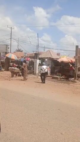 The video shows a street scene in what appears to be a market area. There is a dirt road with a few parked cars and several yellow auto-rickshaws. People are walking around and some are gathered near stalls or shops. There are several buildings visible, some with corrugated metal roofs. There are power lines running overhead and the sky is partly cloudy. Overall, it appears to be a typical bustling market scene in a developing country.