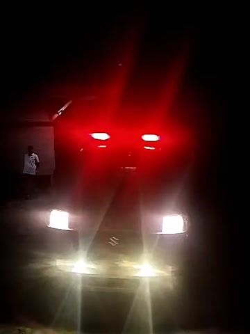 A car, likely a Suzuki, is parked at night with its headlights and red emergency lights flashing. A person is standing near a building to the left of the car. The area is dark except for the light emitted by the car.