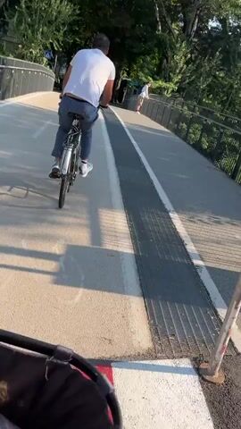 A man cycles on a path next to a river. Trees line the riverbank, and mountains are visible in the distance. The sun is shining.