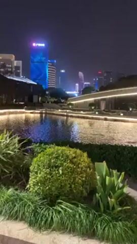 The video shows a nighttime view of a modern urban landscape with a body of water in the foreground. The water is illuminated by lights along its edges, creating a bright reflection. Behind the water is a walkway or platform with architectural details and seating areas. In the background, there is a skyline of tall buildings, some with distinct shapes and illuminated with various colors. The scene appears tranquil and well-maintained, suggesting a public space within a city. The camera slowly pans from left to right, showcasing the urban landscape.