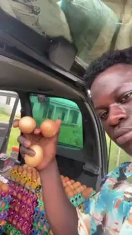 A man is holding three eggs in his hand, displaying them to the camera. He is standing near the back of a vehicle with the trunk open. The trunk is full of egg cartons stacked high.  The egg cartons are various colors. The man is wearing a colorful shirt and is looking directly at the camera with a slightly serious expression. The background shows a building and some greenery.