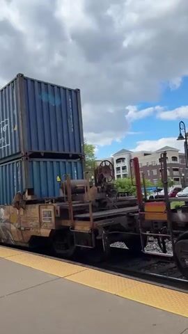 A video shows a train passing by a yellow-tiled platform. The train cars are tan with graffiti and carry two stacked shipping containers each. The containers are blue and red, with shipping information and company names printed on their sides. The sky is cloudy.