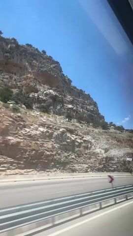 The video shows a view from a moving vehicle, likely a bus or car, looking out a window. The scene is a mountainous landscape with a clear blue sky. A highway runs through the scene, with guardrails visible along the side. A white truck with a blue tank trailer is driving on the highway. The mountains are rocky and steep, with sparse vegetation. The sun is shining brightly, casting shadows.