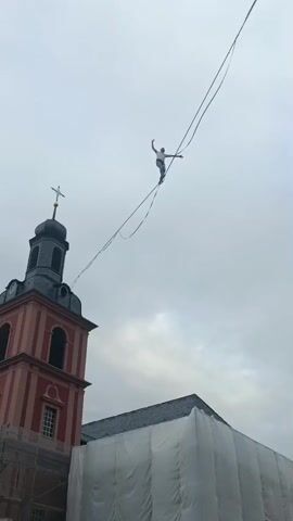A man is walking on a tightrope high in the air. He is dressed in a white shirt and black pants. He is balancing with his arms outstretched. In the background, there is a tall red and gray church tower with a cross on top. Next to the church, there is a building covered in white tarps. The sky is cloudy and gray. The tightrope is strung between two unseen points.