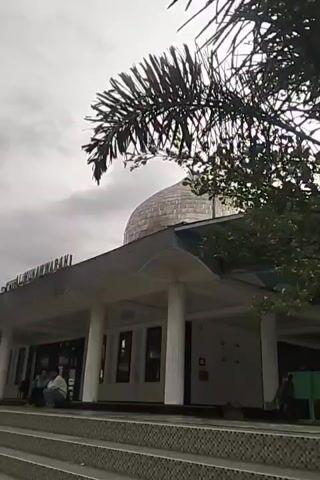 The media file shows an exterior view of a mosque in Blang Bebangka, Indonesia. In the foreground, a set of wide concrete stairs leads up to the mosque's entrance. The mosque features a large, silver dome and a covered walkway supported by white pillars. The walkway has a dark, wavy trim along its edge. Green text on the building reads "AL IMAMUN WARASAH".

Two individuals are seated on the steps near the mosque entrance. Further back, to the left, a bridge crosses over a river. The river is flowing with some rapids. Another, smaller bridge is visible beneath the larger one. To the right of the mosque, a bit of a green structure is visible.

In the upper right portion of the frame, palm fronds hang over the scene. The sky is overcast with grey clouds, suggesting it may be an early morning, late afternoon, or simply a cloudy day. The overall mood appears peaceful and serene.