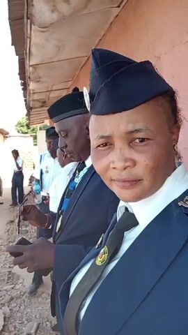 The video shows a group of people, mostly in uniform, standing outdoors. The primary focus is on a woman in the foreground wearing a dark blue uniform with a matching hat and a patterned tie. She is smiling and looking directly at the camera. Behind her and slightly to the side, another man in a similar uniform and hat, but also wearing sunglasses, is visible. Further back, other individuals in uniform are partially visible.  A car is parked in the background. The setting appears to be outside a building with a red wall. The ground is a mixture of dirt and gravel. The camera appears to be handheld, with the woman possibly taking a selfie or being filmed by someone close by.