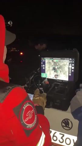 A group of people are gathered around a vehicle at night. One person, wearing a red hooded jacket with a patch on the sleeve, is looking at a tablet and a larger screen. The larger screen displays a thermal image with several markers. Another person, wearing a black hoodie, is standing behind the tablet. In the background, other individuals and vehicles are visible in the darkness. The overall scene suggests some sort of operation or activity taking place outdoors at night.