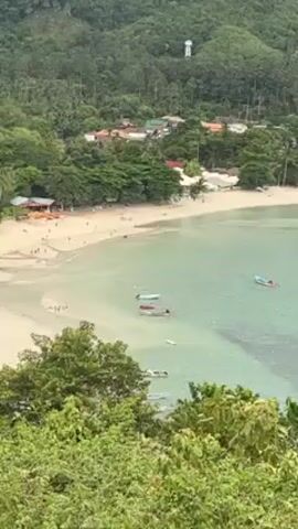 The video shows an aerial view of a tropical beach and coastal village. The beach is sandy and curves gently along a bay. People are visible on the beach, walking and relaxing. Several small boats are floating in the turquoise water near the shore. Lush green vegetation, including trees and bushes, cover the surrounding hillsides. Buildings, mostly low-rise and colored white and brown, are nestled among the trees near the beach.  A tall white structure, possibly a water tower, is visible on one of the hills in the background. The overall scene is bright and sunny.