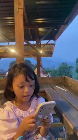 The video shows a young girl sitting at a wooden table outside, looking at a smartphone. The camera pans from the girl to reveal a view of trees, hills, and rice paddies in the distance, under a cloudy blue sky. The scene is partially covered by a wooden roof structure with hanging lights. Other people can be seen in the background.