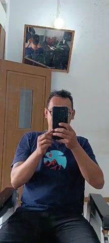 A man wearing glasses and a dark blue t-shirt with a graphic print is sitting in a chair and holding a smartphone to take a selfie. He is touching his hair with his left hand and gesturing with his right hand. In the background, there is a mirror reflecting the man and a barbershop setting with tools and products. A wooden door is also visible to the left of the man. The room appears to be well-lit.