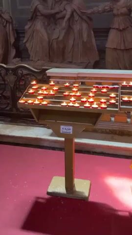 The video shows the interior of a church or religious building. In the foreground is a stand holding many lit votive candles. Behind the candles is a wooden sculpture of several figures in distress, apparently mourning a body laid out. Above the sculpture hangs a large framed painting depicting a religious scene with many figures. The overall tone of the video is solemn and reverent.