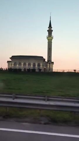 A tall, slender minaret of a mosque stands proudly against a pale sky, its pointed spire reaching upwards. The mosque itself is a stately, rectangular building, illuminated by rows of glowing windows. The structure sits atop a gently sloping, verdant hill. In the foreground, a highway barrier, a series of metal rails, separates the viewer from the peaceful scene. The time of day appears to be either dawn or dusk, as the sky transitions through soft shades of pink and orange.