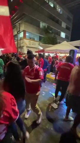 The video shows a street party at night. A crowd of people, many wearing red shirts, are dancing and celebrating. Some are holding flags, and music is playing. The scene is lit by streetlights and colored lighting. There are tents and buildings visible in the background. The atmosphere appears festive and energetic. Debris litters the ground.