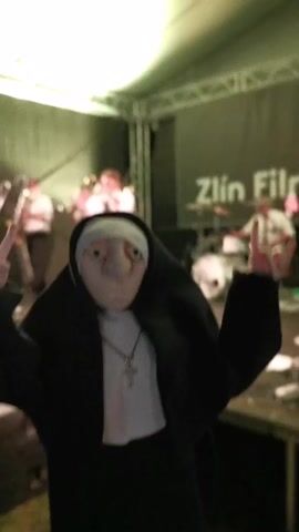 The video shows a person wearing a nun mask dancing. The person is wearing a black nun habit and a cross necklace. They are making the "rock on" hand gesture with both hands. Behind the masked person, there is a band playing music on a stage. The stage has green lighting and a banner that says "Zlín Film". There are people dancing and watching the band in the background. The camera is slightly shaky.