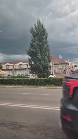 The video shows a view from inside a moving vehicle. In the foreground, the side of a dark gray SUV is visible. The background depicts a street scene with several buildings, trees, and people. The sky is overcast with dark, stormy clouds. There is a supermarket with a red sign and a billboard visible on one of the buildings. Several cars are parked and driving on the street. A few people are walking across a crosswalk, and one person is riding a scooter. Traffic lights are visible, with some showing red. The overall impression is a cloudy, possibly inclement weather day in an urban or suburban setting.
