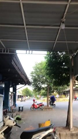 The video shows a view from under a covered structure looking out at an outdoor area. In the foreground, the rear of a white scooter is visible. In the midground, a red scooter with a blue bag on the back is parked. Behind the red scooter, there are trees and a street with some buildings. The sky is visible and appears to be daylight. There is a light fixture hanging from the ceiling of the covered structure. The ground is paved.