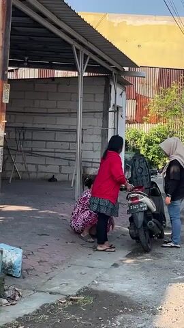 The video shows three women interacting near a parked scooter. One woman, wearing a red hoodie and black leggings, is crouched down near the scooter, seemingly attending to it. Another woman, in a patterned dress, is also crouched beside the scooter, with her back to the camera. A third woman, wearing a hijab, a black top, and blue jeans, stands to the right of the scooter and is looking at the other two women, with her mouth open as if speaking. The scooter's seat is open. The environment appears to be outdoors, possibly in a residential or commercial area, with a building structure in the background.