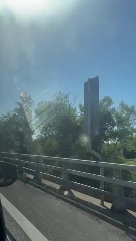 The video shows a view from inside a vehicle looking out a window. The perspective is from the driver's side, with the side mirror visible in the bottom left corner. The vehicle appears to be on a bridge with a concrete barrier and a road. Lush green trees and foliage line the side of the road beyond the bridge. The sky is clear and blue with a bright sun creating lens flare and reflections on the window. The sun is positioned in the upper left quadrant of the frame. There are no people visible in the video, and the focus is on the road, bridge, and surrounding natural environment. The overall impression is a sunny day drive.