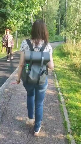 A woman with dark hair walks away from the camera on a paved path. She wears a floral print shirt, blue jeans, and a dark gray backpack. A green water bottle is clipped to the side of her backpack. A second person, wearing a patterned shirt and dark pants, walks on the path to the left of the woman. The path is bordered by green grass and trees. The sun is shining, casting shadows on the path.