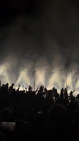 This video captures a vibrant nighttime celebration, likely a festival, in Jávea/Xàbia, Spain. The scene is dominated by a large crowd of silhouetted people, their faces obscured by the darkness, suggesting an event occurring after sunset. They are gathered in an open area, possibly a town square or promenade, with buildings visible in the background.

The main activity appears to be a pyrotechnic display. Streaks of golden sparks erupt in the air, illuminating the night sky and creating a dazzling spectacle. These are accompanied by plumes of smoke, adding to the dramatic atmosphere. Several tall, thin structures topped with spiky elements are dotted throughout the illuminated area, possibly part of the fireworks setup or decorative elements for the festival.

Many individuals in the crowd are holding up their phones, presumably to capture the event, a common behavior at modern celebrations. Some are raising their arms in the air, indicative of excitement and engagement with the festivities. The overall mood seems to be one of joy and exhilaration, with the visual elements of light and smoke contributing to a sense of awe and wonder. There is no visible text within the media file.