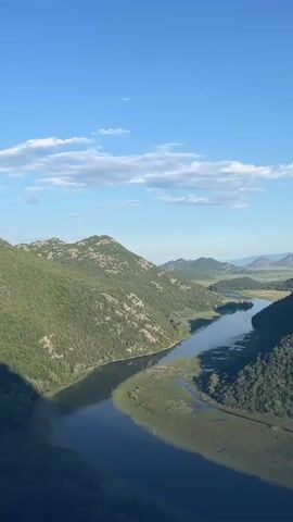 The video shows a drone view over a winding river surrounded by lush green hills. The sky is blue with scattered white clouds. The river appears to be flowing through a valley, with vegetation growing along its banks. In the distance, there are more hills and mountains. The drone moves slowly, offering a sweeping panorama of the landscape. At one point, a small boat can be seen on the river, moving away from the camera. The sun appears to be shining, casting shadows on the hills. The overall impression is one of natural beauty and tranquility.