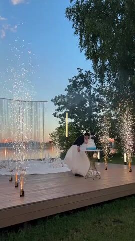 A wedding couple stands on a wooden deck in front of a tiered wedding cake. The bride is in a white gown, and the groom is in a dark suit. They are embracing and appear to be kissing. Fireworks are shooting up around them. The sky is a pale blue, transitioning to a soft orange near the horizon. Trees and foliage are visible in the background. The scene appears to be outdoors during twilight.