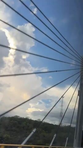 A camera moves past the cables and white tower of a cable-stayed bridge. Through the gaps in the bridge structure, a green, forested hill is visible. A tall, slender communication tower stands atop the hill. The sky is partly cloudy with patches of blue and white clouds, and sunlight is visible on the left side of the frame.