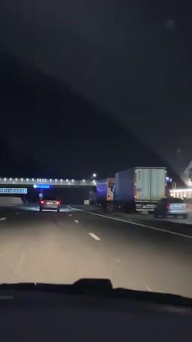 The video is filmed from the perspective of a person in a car at night, driving on a highway. The road ahead is illuminated by streetlights and the headlights of other vehicles. In the distance, a pedestrian bridge with blue lights spans across the highway. To the right of the highway, there is a truck stop or gas station with a red "ГАЗ" sign. Several trucks are parked along the right side of the highway. A few cars are visible on the road ahead, including a silver car in the left lane. The overall environment is dark, with artificial lighting from the road and nearby businesses.