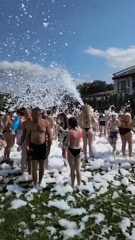 A large group of people are gathered outdoors on a grassy area, covered in a thick layer of white foam. A machine is spraying a continuous stream of foam into the air from the left side of the frame, creating a dense cloud of foam that obscures the sky in that area. The people are mostly wearing swimwear, with some in bikinis and swim trunks. They appear to be enjoying themselves, with many smiling and interacting with each other. The environment is sunny with a clear blue sky and some scattered clouds. There are trees and buildings visible in the background.