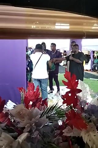 The camera is angled upwards, positioned behind a curtain of flowers featuring red, white, and purple blooms.  Focusing through this floral veil, several individuals are visible. One person wears a white shirt and blue jeans, while others are dressed in darker clothing. They appear to be standing or milling around an area that is decorated with a purple backdrop. The scene seems to capture an event, possibly a gathering or celebration, viewed from a somewhat obscured vantage point.