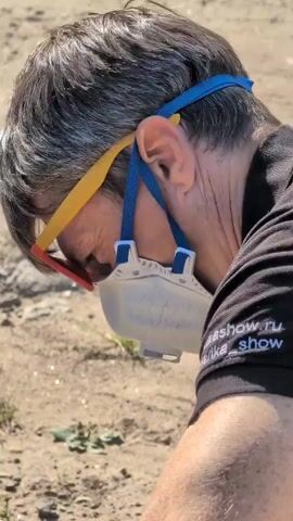 The video shows a close-up of a person wearing sunglasses, a dust mask, and a black shirt with yellow and blue straps. The person's head is bent down, and their gloved hands are visible in the lower part of the frame, interacting with something off-camera. The environment appears to be outdoors, with dirt, small plants, and scattered rocks visible. Text is visible on the person's shirt, reading "akeshow.ru" and "harika_show."