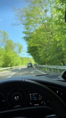 A first-person perspective video shows a driver on a rural road in what appears to be a car. The camera is positioned behind the steering wheel, capturing the view of the road ahead through the windshield. On the dashboard, a digital display shows the car's speed fluctuating around 63 km/h, along with the date "17.05.2026," a temperature reading of 39°C, and a voltage reading of 13.6V. The car's driving mode is set to "NORMAL." The road is flanked by green trees on both sides. The sky is blue and clear. In the distance, two cars are visible on the road ahead. The overall environment suggests a sunny day in a forested or rural area. The video quality is moderately sharp, with the road and surrounding greenery clearly visible.