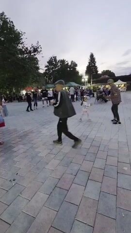The video shows an outdoor public space filled with people, a stage, a bounce house, and other amenities. A small girl in a light jacket and pink pants runs across a brick-paved area toward a stage.  A person in dark clothing jogs behind her. To the right of the stage, a bounce house stands. The background reveals people gathered under small tents and seated at tables. Others are standing and milling around.  A band performs on stage. The sky is overcast.  The scene appears to be a local festival or event. The video also features two other men, one middle-aged, one young, casually walking around the space.