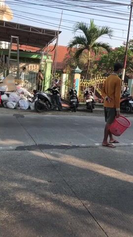 The video depicts an outdoor scene with a paved road in the foreground. A person wearing a yellow shirt, grey shorts, and a mask walks from right to left, carrying a red basket. To the left of the road, there is a pile of white bags, some of which appear to be trash bags, and several motorcycles are parked. Behind the motorcycles, there is a fence and a building with a red roof and a green gate. Palm trees and other vegetation are visible in the background. The lighting suggests it is daytime, with shadows cast on the road.