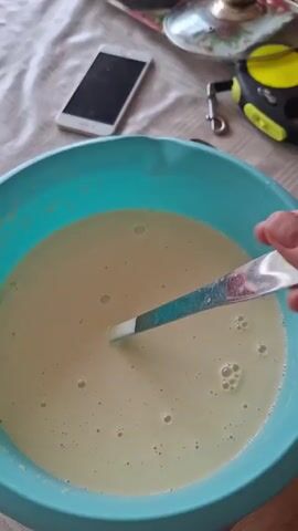 The video shows a close-up of a person's hand holding a ladle full of a pale yellow liquid. The ladle is being lifted out of a large, light blue plastic bowl. The liquid appears to be a batter or dough. In the background, on a table covered with a patterned cloth, is a white smartphone, a glass lid with a decorative handle, and a yellow and black retractable tape measure. The lighting suggests an indoor environment, possibly a kitchen. The action is focused on the process of scooping the liquid.