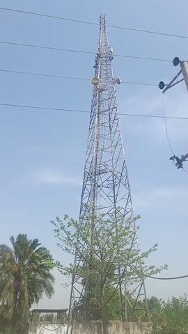 The video shows a tall telecommunications tower made of metal lattice. The tower has several cylindrical objects attached to it, likely antennas or other communication equipment. It is surrounded by trees and a concrete utility pole carrying electrical wires and a transformer. The sky is mostly clear and blue. The camera appears to be moving, likely from a passing vehicle.