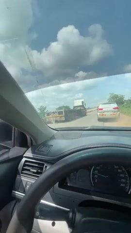 A first-person perspective video shows a view from inside a car, looking out the windshield and side window. The car is on a road, possibly in traffic, as several vehicles are visible ahead. In the foreground, the steering wheel and dashboard of the car are in focus. Outside, there is a large orange truck in the middle distance, and a white car further ahead. The sky is cloudy with bright patches of blue. Dust appears to be kicked up on the road, suggesting the vehicles are moving or have recently moved. The road appears to be paved but has some rough patches. Trees and vegetation are visible along the side of the road. The overall impression is of a sunny day with some road travel activity.