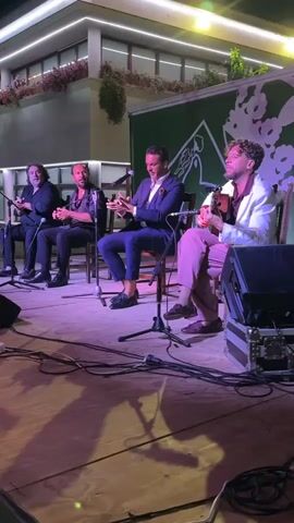 Four men are seated on chairs on a wooden stage, performing music. The man on the far left is seated and playing an instrument, possibly a guitar, with a dark shirt. The second man from the left is seated and clapping his hands, wearing a dark shirt and pants. The third man from the left is seated, wearing a dark suit and blue pants, and clapping his hands. The man on the far right is seated, wearing a white jacket and pink pants, playing a guitar. There are microphones in front of each performer. A green banner with a white design is behind them, and above them is a building with lights and plants. The floor is made of wood, and there are cables and audio equipment in the foreground. The lighting is dim and purple.